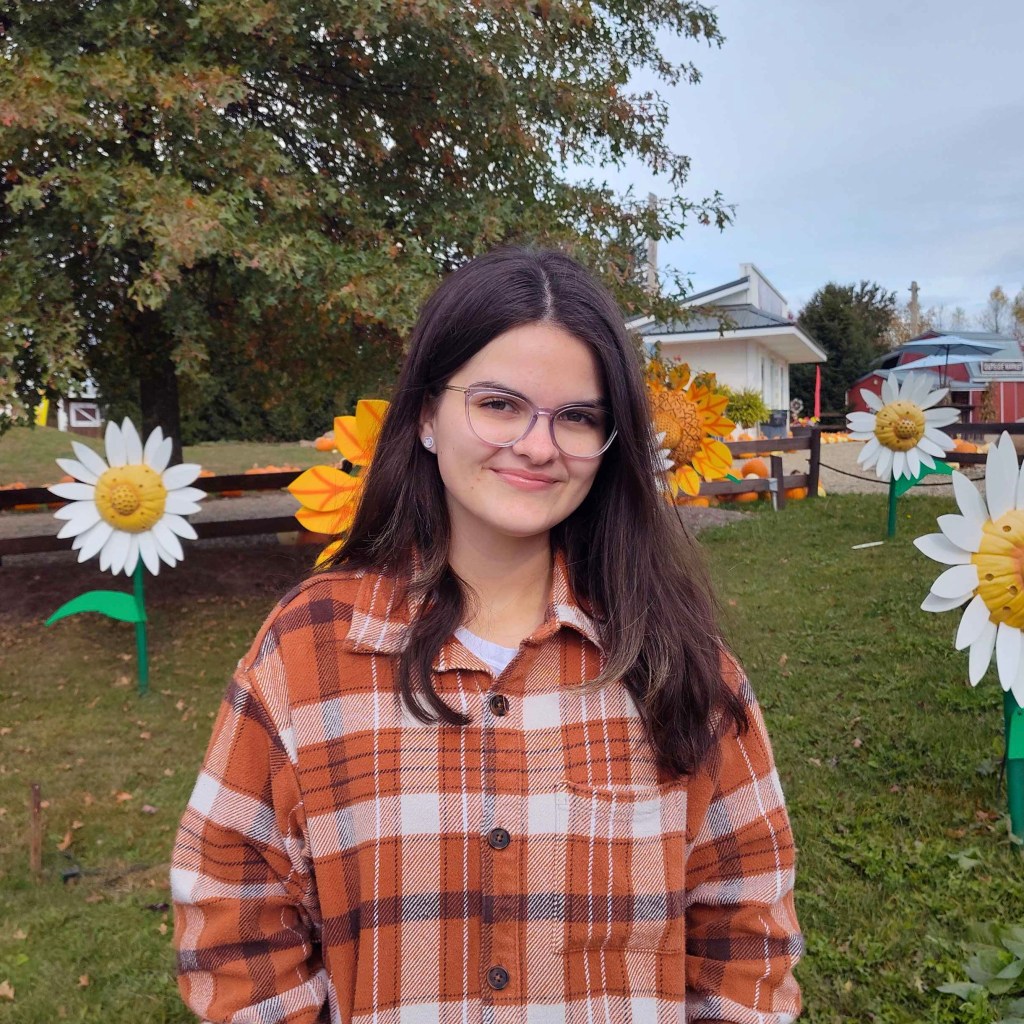 A young woman with long dark hair and glasses smiles at the camera from a grassy field filled with artificial sunflowers.