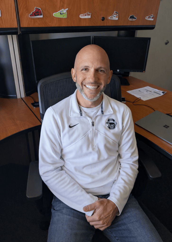 Photo of a smiling bald, bearded man wearing a Penn State pullover, sitting at a desk. 