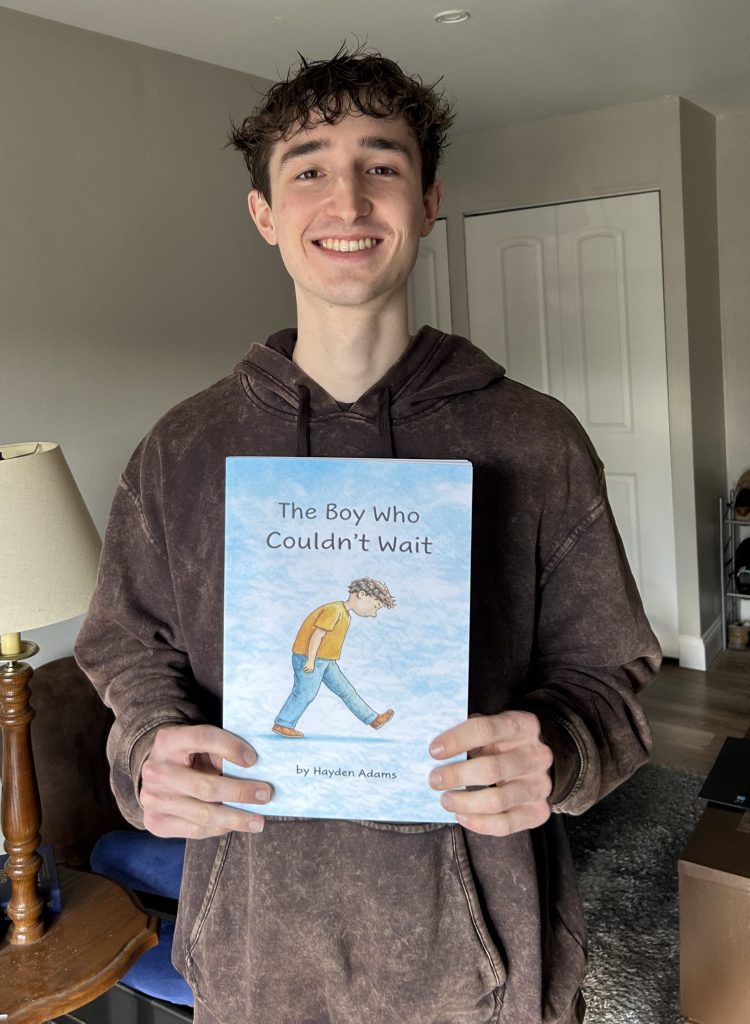 Young man wearing a brown hoodie holding a large hardcover children's book with a watercolor illustration. 